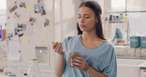 Pretty hispanic girl cheeky face eating chocolate spread from jar using spoon wearing pajamas at home by window