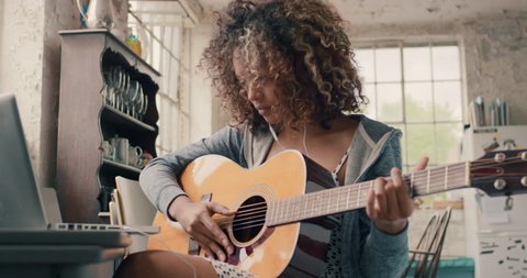 Attractive curly haired mixed race young girl sitting on wooden chair at a window wearing a grey hoodie concentrating focused learning to play guitar using laptop computer at home