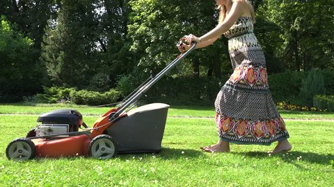 barefoot young woman in long summer dress cut green grass in own home yard with lawn mower. Handheld shot. 4K UHD video clip.