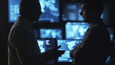 Two male employees are having a conversation in a dark monitoring room filled with display screens. Shot on RED Cinema Camera in 4K (UHD).