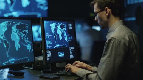 IT professional programmer in glasses is working on computer in cyber security center filled with display screens. Shot on RED Cinema Camera in 4K (UHD).