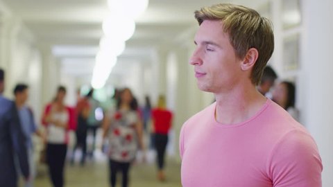 Portrait of a happy male caucasian student standing in a busy hallway with other students and teachers walking in the background. In slow motion.