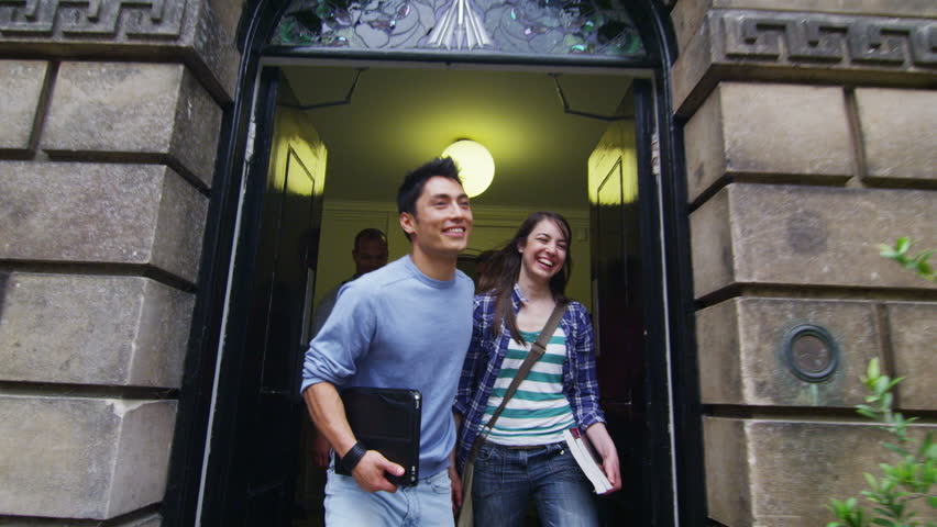 Large group of students of mixed ethnicity leaving through the main door of their historic university or college building. In slow motion.
