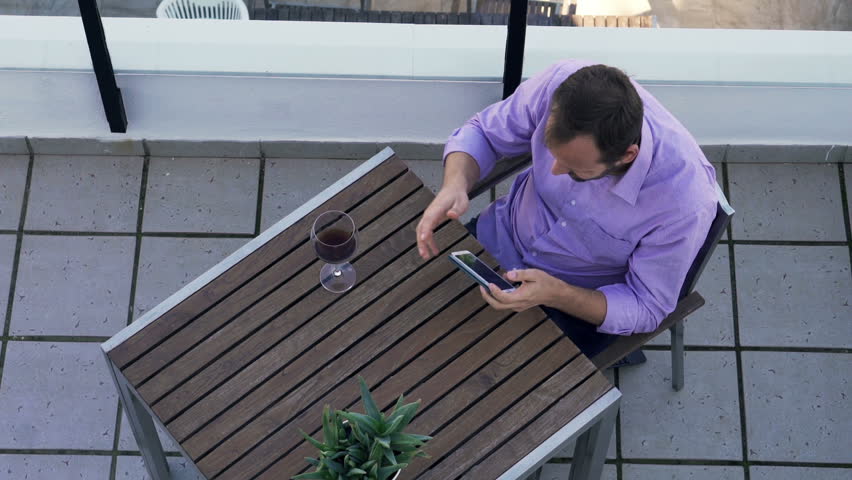 Young man playing game on smartphone sitting by table on terrace, top view
