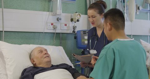 4k / Ultra HD version Nurses attend to an elderly male patient, checking his blood pressure and chatting with him. In the background a nurse and a specialist discuss another patient. Shot on RED Epic