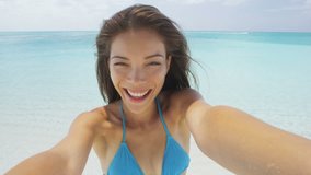 Gorgeous cheerful young woman taking selfie on beach. Portrait of carefree girl enjoying her summer vacation. Happy female is in blue bikini top on perfect white sand beach on travel holidays.
