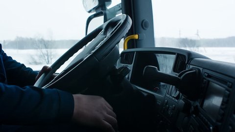 The driver starts a truck, then removes the handbrake and starts to move in a comfortable cabin. He goes on snow-covered road. Worker turns steering wheel of truck. Snow