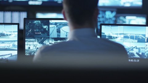 Security officer is working on a computer in a dark monitoring room filled with display screens. Shot on RED Cinema Camera in 4K (UHD).