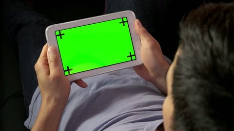 Asian man touching ipad computer monitor on couch at home, people using digital tablet with green screen, lying on sofa, relaxing. Wi-fi technology for internet and email, lifestyle
