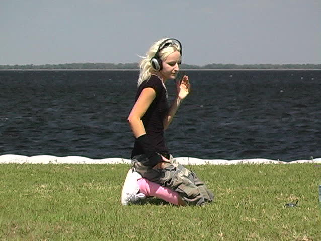 A beautiful, hip blonde on her knees in the grass at a lakefront moves to the beat of the music in her portable mp3 player with headphones.