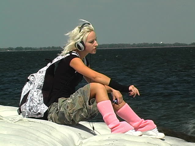 A beautiful, hip blonde sitting on a lakeshore enjoying the music from her portable mp3 player with headphones.