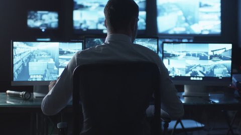Security officer is drinking coffee while working on a computer in a dark monitoring room filled with display screens. Shot on RED Cinema Camera in 4K (UHD).