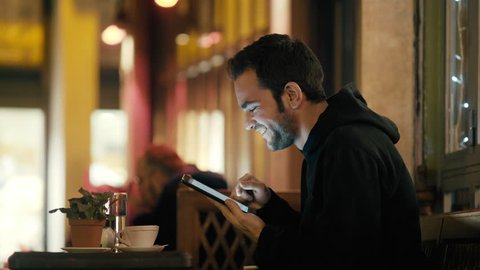 4K Young man sitting in night cafe/bistro browsing internet social media tablet, outside at winter at night after work drinking coffee and browsing his sites and social media on his tablet.