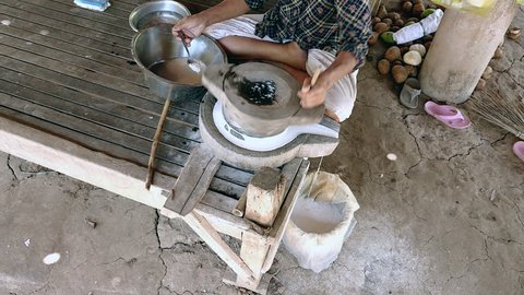 Upper view on woman sat cross-legged using hand-turned millstone to grind wet rice for making soaked rice flour