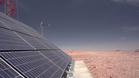 Panning shot of a solar panel (and a small wind turbine) install in the top a mountain. The background is the Atacama desert in Chile