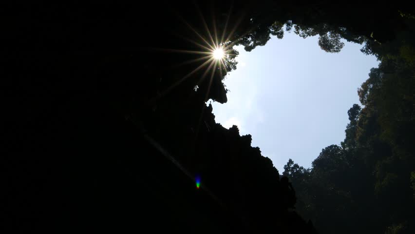 Blue sky sunbeam star through cave ceiling opening, look up, view surrounded by cave walls, dark foliage and tree leaves on sides. POV camera slowly move, nice changes of the sun rays and brightness