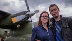 Couple take selfie photo in front of big army aircraft. Young man and woman stand in front of an aircraft with big propellers on a wing. Smiling in camera. Portrait shot.