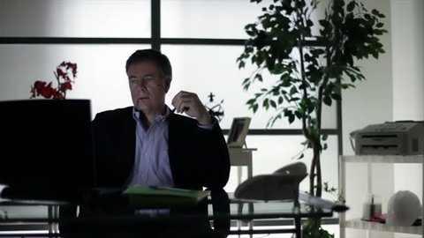 A businessman sitting in his dimly lit office working at his computer contemplates a problem he is trying to address.