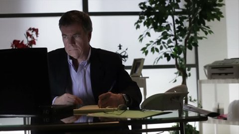 A businessman sitting in his dimly lit office writing something on a tablet from information he has on his laptop.
