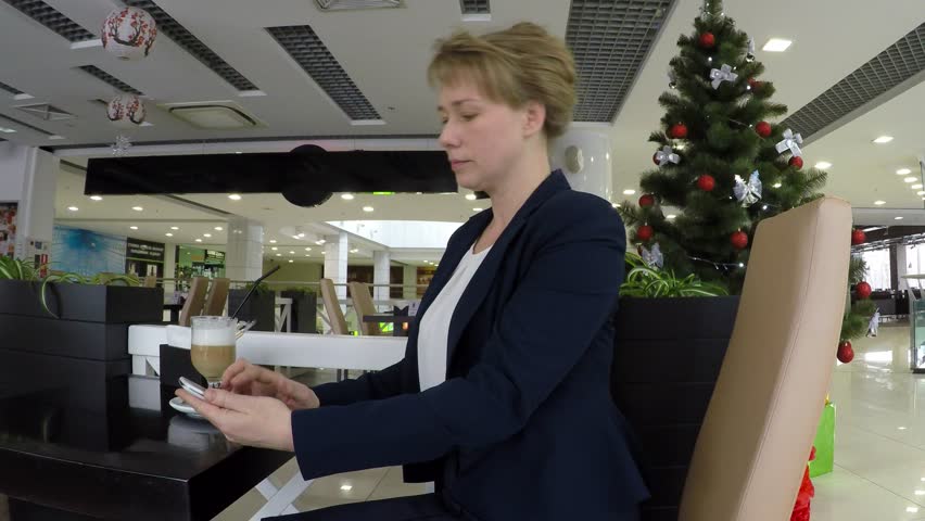 In the background a Christmas tree. On the table is a cappuccino. Young beautiful woman sad on the phone.