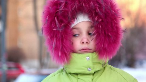 Little girl in big red fur hood buttoned at collar, look at camera and rolls eyes