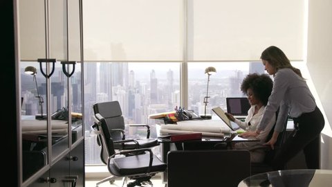 Two architects in modern office building, sitting on desk with blueprints and housing projects. The women hold a tablet and surf the web. They smile and talk each other. Wide shot
