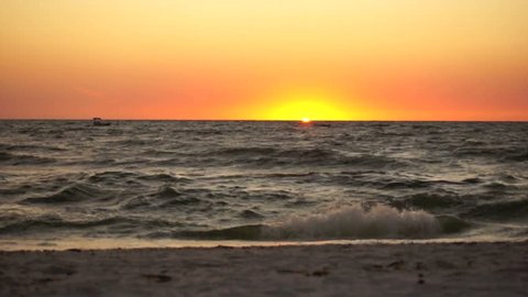 A boat slowly crosses the scene as the sun dips below the horizon with waves crashing in the foreground of this sunset clip of bonita beach in bonita springs florida.