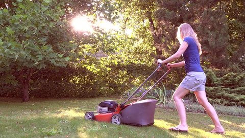 Blond worker woman mowing the grass during the beautiful evening sunlight in yard. Static shot. 4K