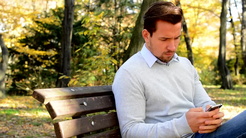 Young man sits on bench in the park, thinks about something and works  on the phone
