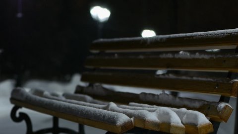 Snow falls on wooden bench in park