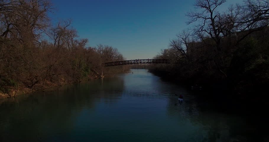 The camera starts low and then gains altitude over the Barton Springs pedestrian bridge in Austin, Texas.  Pedestrians and runners cross the bridge as downtown reveals itself in the background.