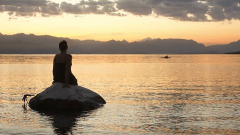 Successful young woman sitting on a rock enjoying the beautiful sunset at the lake and mountains