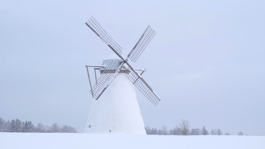 The non-fucntional windmill in snow. The snow covering the entire lawn and the trees behind with snow