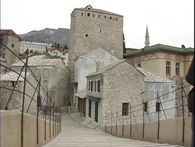 A couple walks down a flight of stairs, toward old stone buildings in Bosnia.