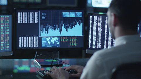Man is working on a computer with data and graphs in a dark office filled with display screens. Shot on RED Cinema Camera in 4K (UHD).