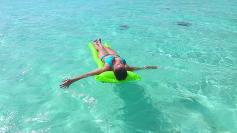 SLOW MOTION: Happy young female in bikini laying and sun tanning on inflatable airbed mattress floating on water surface in beautiful emerald lagoon in Bora Bora island resort