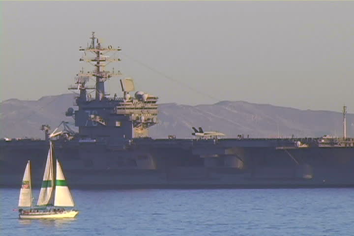A sailboat is dwarfed by an huge aircraft carrier.