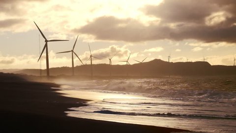 Wind turbine power generators silhouettes at stormy ocean coastline at sunset. Alternative renewable energy production in Philippines