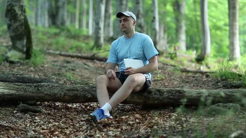 Adult man with white Tablet PC sits on a fallen tree in the wood and listens to music. Music lover. Male with tablet computer. Sportsman with Tablet PC in the forest