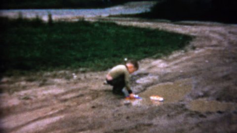 STEEP FALLS, MAINE 1962: Boy playing with toy boats in sad muddy driveway puddle.