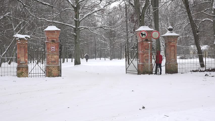 Man with warm clothes enter snowy winter park through retro brick gate entrance and snow falling. Static shot. 4K