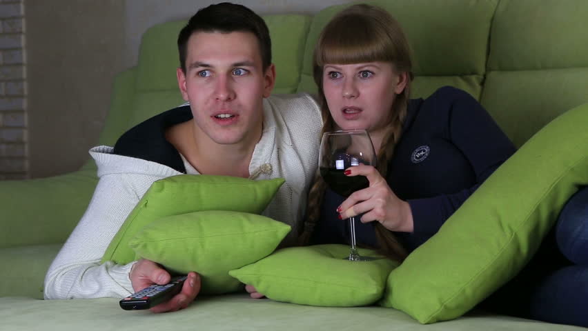 Frightened young couple with glass of wine watching the horrors on television.