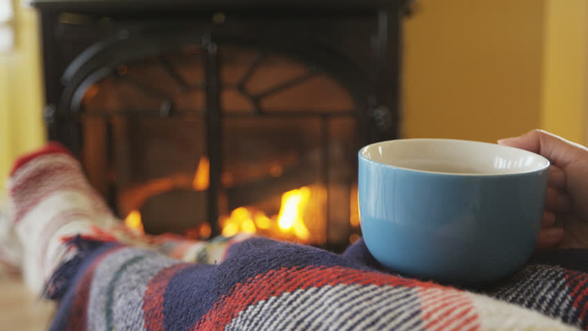 Woman drinking coffee cup nearby fireplace. Girl is covering herself with blanket while having hot drinking getting warm in her living room. She is warming herself in front of burning stove in winter.