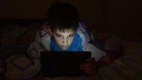 Young boy plays video games in bed covered with blanket at sleeping time in dark bedroom with lights turned off