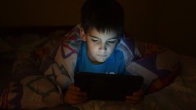 Young boy plays video games in bed covered with blanket at sleeping time in dark bedroom with lights turned off