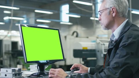 Senior engineer in glasses is working on a desktop computer with a green screen on monitor in a factory. Shot on RED Cinema Camera in 4K (UHD).
