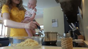 Babysitter holding infant baby in hands take boiled spaghetti from pot to glass dish in kitchen. Housewife multitasking at home. Vapor steam rise. Static shot. 4K UHD video clip.