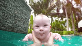 Happy. smiling baby learning to swim with Mommy in an adult sized pool with a backdrop of beautiful palm trees and gardens. UltraHD video
