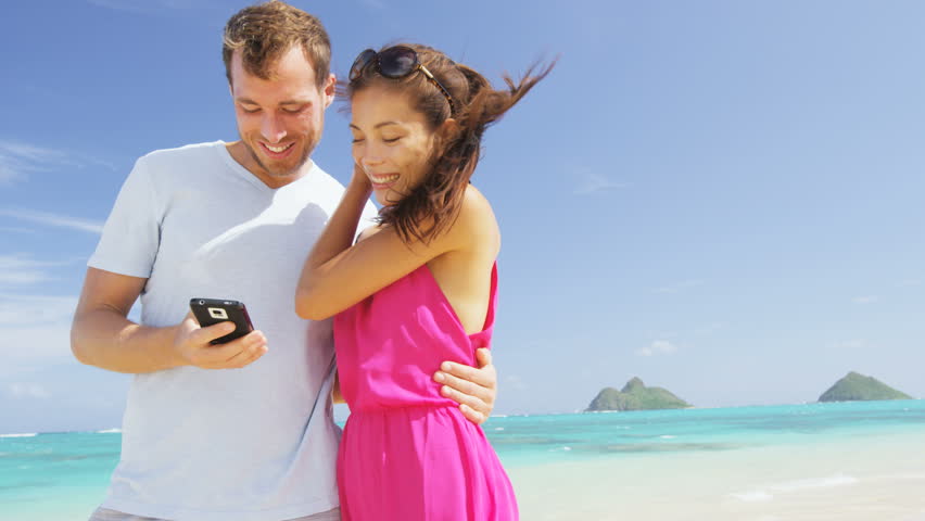 Beach couple on vacation taking smart phone selfie photograph using smartphone camera. Romantic couple in love on honeymoon, Oahu, Hawaii, USA with Mokulua Islands. RED EPIC SLOWMO.