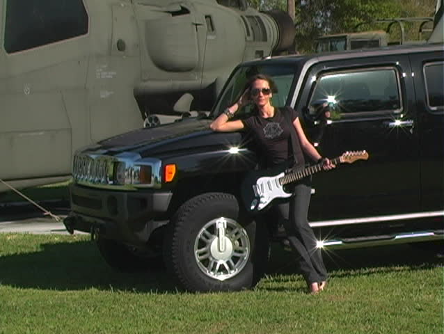 A beautiful brunette with her electric guitar in front of her black SUV, parked in front of a military combat helicopter.  See additional clips from alternate angles to form a series.